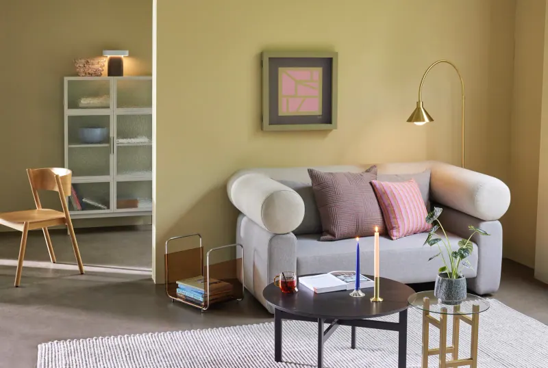 Wohnzimmer mit Möbeln und Leuchten von Hübsch: graues Sofa mit rosa Kissen, Beistelltische und Stehleuchte vor einer gelben Wand