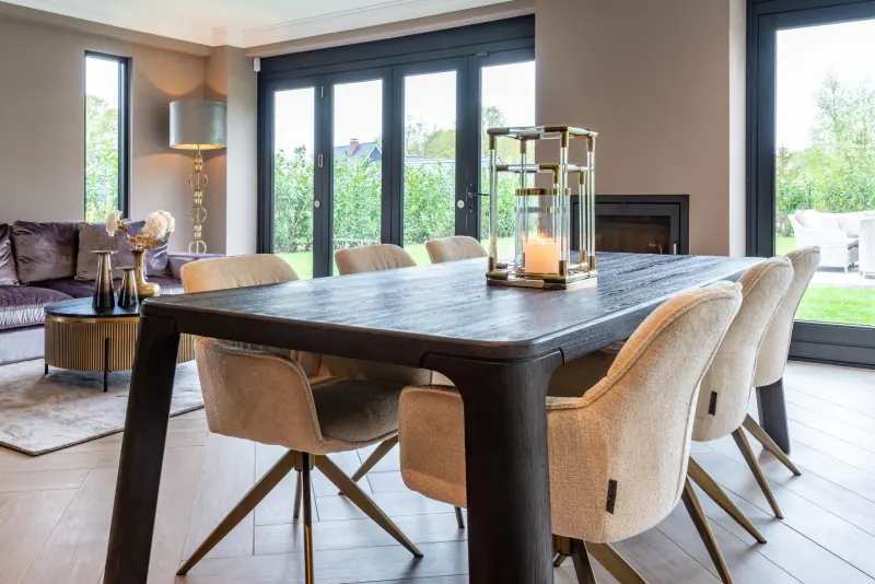 Dunkler Esstisch mit hellen Polsterstühlen und einem goldenen Windlicht von Richmond Interiors vor großen Fenstern