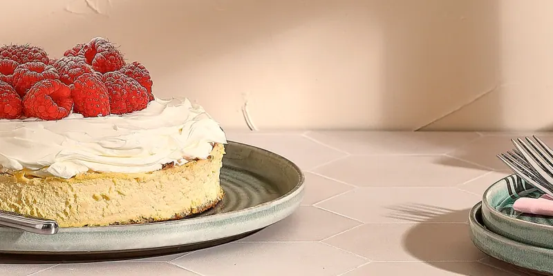 Käsekuchen mit Himbeeren auf einem grünen Teller mit reaktiver Glasur der Salt&Pepper-Serie Cameo, daneben gestapelte Teller und Gabeln