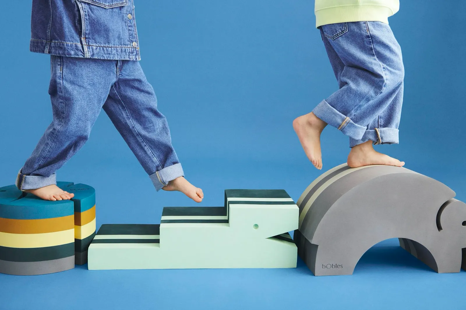 Zwei Kinder steigen über Tumbelelemente von bObles in Mint und Grau vor blauem Hintergrund